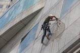 A person is suspended on the side of a modern glass building, wearing safety gear and a helmet while performing maintenance or cleaning tasks. The harness is securely attached, and they are equipped with tools for the job. The reflection of the person is visible on the glass surface.
