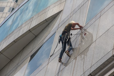 A person is suspended on the side of a modern glass building, wearing safety gear and a helmet while performing maintenance or cleaning tasks. The harness is securely attached, and they are equipped with tools for the job. The reflection of the person is visible on the glass surface.