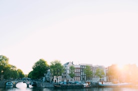 A tranquil canal scene with characteristic Dutch architecture lining the waterway. Trees with lush green foliage and boats gently floating on the canal add charm to the serene setting. The warm glow of sunlight creates a pleasant atmosphere.