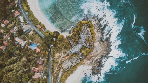 An aerial view captures a coastal area with clear turquoise waters hitting the rocky shoreline. A road curves alongside densely packed houses with red and gray roofs surrounded by lush green vegetation. A distinctively shaped, partially constructed building lies near the coastline, appearing abandoned amidst the trees.