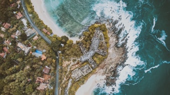 An aerial view captures a coastal area with clear turquoise waters hitting the rocky shoreline. A road curves alongside densely packed houses with red and gray roofs surrounded by lush green vegetation. A distinctively shaped, partially constructed building lies near the coastline, appearing abandoned amidst the trees.