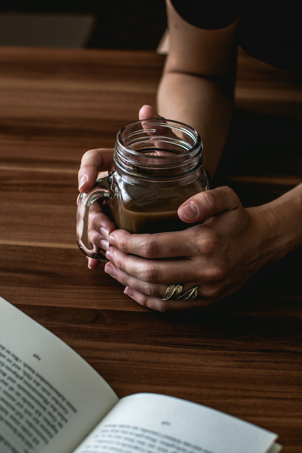 person leaning on table while holding Mason jar mug with brown liquid