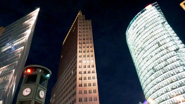 A cityscape featuring three distinctive buildings, including a modern glass structure, a brick skyscraper, and a cylindrical glass tower. There is also a traffic light structure with multiple clocks displaying different times, set against a dark night sky.