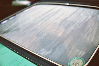 Raindrops scattered across the surface of a car window, creating streaks and patterns. A visible sticker is located in the bottom right corner. The focus highlights the texture of the water droplets against the glass.