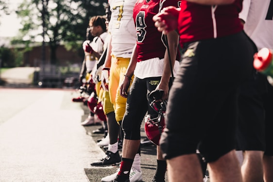 A group of football players stand in a line on the sidelines. They are dressed in colorful uniforms, with one player holding a helmet. The scene takes place on a sunny day, casting shadows on the ground.