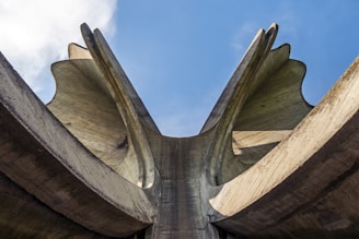 A large, abstract concrete structure with curved, wing-like forms reaching upwards against a blue sky. The monument has smooth surfaces and sharp edges, creating an imposing and dramatic silhouette.