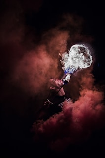 A performer is captured in the act of fire breathing, with flames and smoke artistically enveloping the scene. The background is dark, enhancing the dramatic contrast of the illuminated fire and smoke. The colors range from fiery orange to deep red and the faint silhouette of the individual highlights the intensity of the performance.