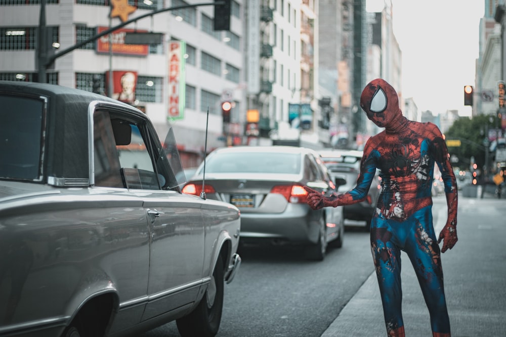 스파이더맨 의상을 입은 남자가 도로에 자동차가 있는 보도에 서 있다.