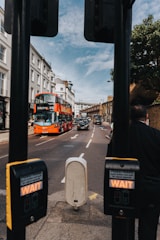 A busy city street scene with a red double-decker bus heading toward Tooting Station. The bus is followed by several black taxis under a partly cloudy sky. Pedestrian crossing buttons are visible in the foreground, with a streetscape of buildings lining both sides of the road.