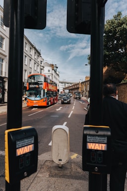 A busy city street scene with a red double-decker bus heading toward Tooting Station. The bus is followed by several black taxis under a partly cloudy sky. Pedestrian crossing buttons are visible in the foreground, with a streetscape of buildings lining both sides of the road.