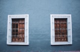 Two adjacent wooden windows with white frames and metal bars are set into a textured blue wall. The design is symmetrical with each window featuring a grid pattern and decorative bars.