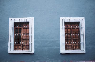 Two adjacent wooden windows with white frames and metal bars are set into a textured blue wall. The design is symmetrical with each window featuring a grid pattern and decorative bars.
