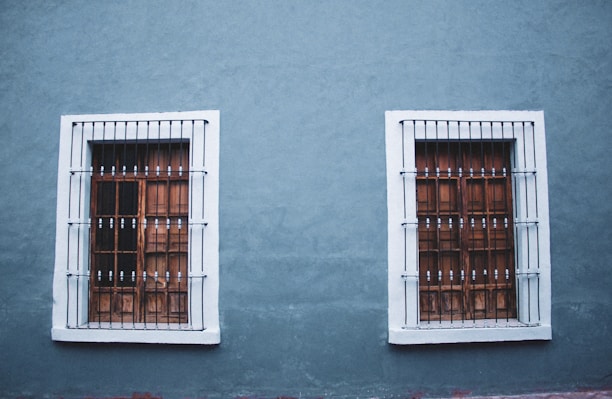 Two adjacent wooden windows with white frames and metal bars are set into a textured blue wall. The design is symmetrical with each window featuring a grid pattern and decorative bars.