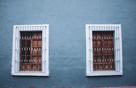 Two adjacent wooden windows with white frames and metal bars are set into a textured blue wall. The design is symmetrical with each window featuring a grid pattern and decorative bars.