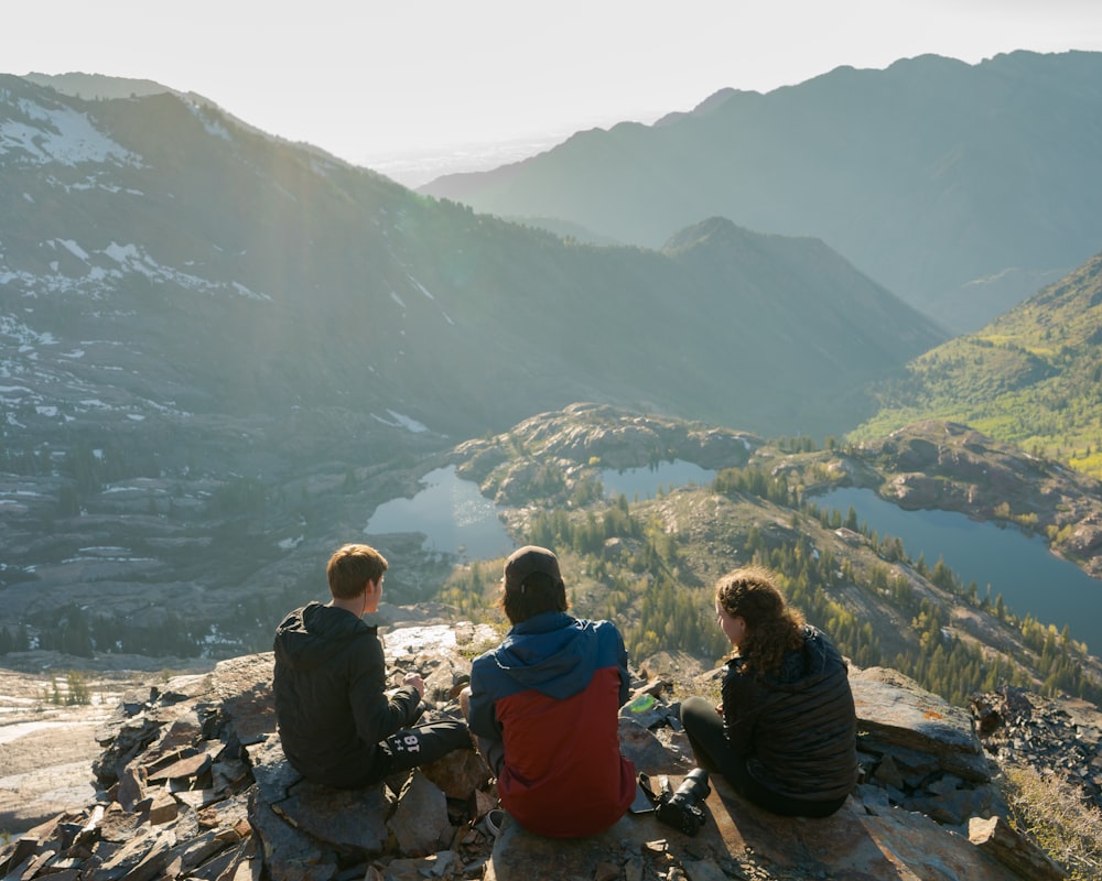 two men and one woman sitting on cliff overlooking lake and mountains during daytime