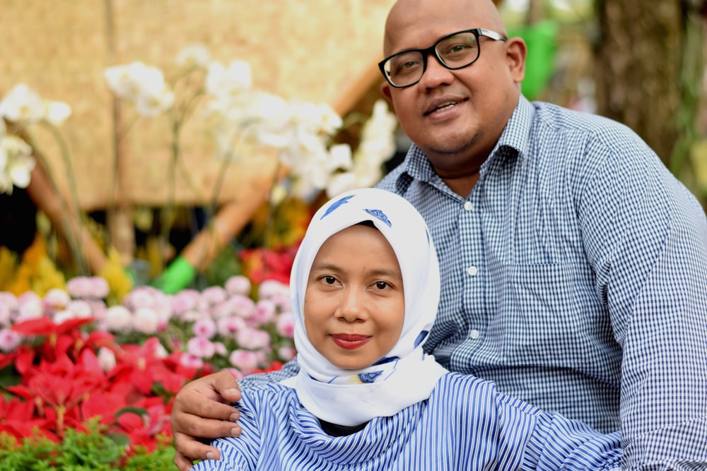 man wearing black framed eyeglasses and woman wearing white hijab taking photo beside flowers