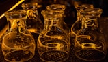 A collection of empty glass jars is placed upside down on a patterned mat. The jars are illuminated by warm, amber lighting, creating reflective patterns on their surfaces. Their transparent nature allows the light and surrounding reflections to be clearly visible, creating a visually striking effect.