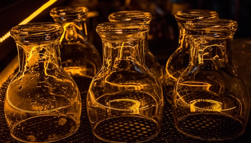 A collection of empty glass jars is placed upside down on a patterned mat. The jars are illuminated by warm, amber lighting, creating reflective patterns on their surfaces. Their transparent nature allows the light and surrounding reflections to be clearly visible, creating a visually striking effect.