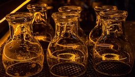 A collection of empty glass jars is placed upside down on a patterned mat. The jars are illuminated by warm, amber lighting, creating reflective patterns on their surfaces. Their transparent nature allows the light and surrounding reflections to be clearly visible, creating a visually striking effect.