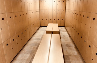 A room filled with wooden lockers lining the walls, creating a narrow passage. The lockers have small square ventilation holes and handles. Two long benches are placed parallel to each other in the center of the room, matching the wood color of the lockers. The floor is tiled with light beige tiles.