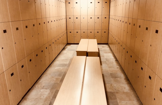 A room filled with wooden lockers lining the walls, creating a narrow passage. The lockers have small square ventilation holes and handles. Two long benches are placed parallel to each other in the center of the room, matching the wood color of the lockers. The floor is tiled with light beige tiles.