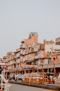 An urban street scene featuring a row of weathered, peach-colored buildings with various signs in an Indian script. The buildings appear densely packed, showcasing distinct architectural features typical of an older city. There is a busy street scene with people riding scooters and auto rickshaws amongst cars as a form of daily transport.