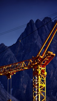 A bright yellow construction crane stands prominently against a backdrop of rugged, dark mountain peaks. The crane's lattice structure is detailed, with cables running from its arm. The lighting casts a dramatic contrast between the warm tones of the crane and the cool, shadowed mountain behind it.