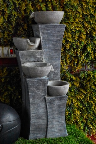 A decorative stone water fountain with multiple tiers is set against a lush green wall covered with artificial or real foliage. The fountain consists of four cascading stone basins supported by a textured stone structure, and is placed on a patch of artificial grass. Nearby, there is a black spherical object, possibly a speaker or ornament. Power outlets are visible in the background.