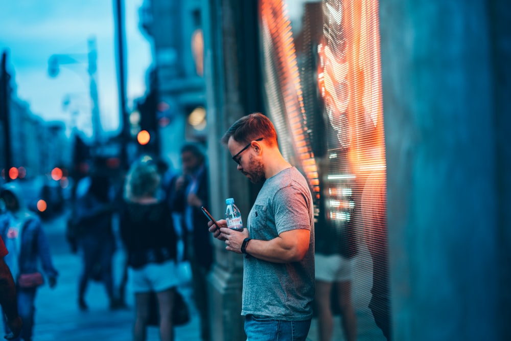 man standing beside wall holding smartphone and water bottle