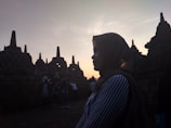 A silhouette of a person wearing a hijab is seen in front of the ancient stupas at Borobudur Temple during sunset, with the sky glowing in soft hues. The scene captures a peaceful moment as other visitors can be seen in the background exploring the site.