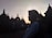 A silhouette of a person wearing a hijab is seen in front of the ancient stupas at Borobudur Temple during sunset, with the sky glowing in soft hues. The scene captures a peaceful moment as other visitors can be seen in the background exploring the site.
