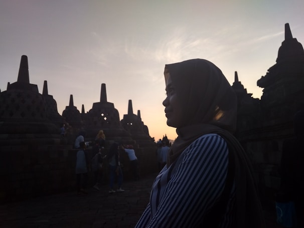 A silhouette of a person wearing a hijab is seen in front of the ancient stupas at Borobudur Temple during sunset, with the sky glowing in soft hues. The scene captures a peaceful moment as other visitors can be seen in the background exploring the site.