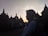 A silhouette of a person wearing a hijab is seen in front of the ancient stupas at Borobudur Temple during sunset, with the sky glowing in soft hues. The scene captures a peaceful moment as other visitors can be seen in the background exploring the site.