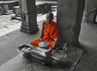 A monk wearing an orange robe is sitting against a stone column in a serene, ancient stone structure. In front of him, there is a mat with a donation box, a bowl, and a plastic bottle. The surroundings are made of stone with carved details.