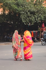 Two people wearing colorful sarees walk arm in arm across a street. They are surrounded by greenery, and there are motorcycles parked on the side. Market stalls in the background display various fabrics and items.