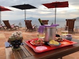 A scenic table setup on a patio with a view of the sea and distant mountains. The table features a wine cooler with a bottle and a purple napkin, two glasses of white wine, a tray with appetizers including chips and bruschetta, and a decorative ceramic container. Wooden deck chairs with colorful cushions are visible in the background under large umbrellas.