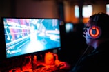 A person wearing headphones is sitting in front of a computer monitor. The screen displays a vibrant, illuminated video game scene, while the person appears focused on gaming. The ambient lighting is dark with red and blue tones, and a backlight from the monitor highlights the headphones.