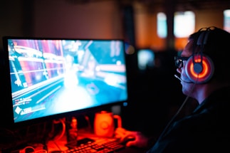 A person wearing headphones is sitting in front of a computer monitor. The screen displays a vibrant, illuminated video game scene, while the person appears focused on gaming. The ambient lighting is dark with red and blue tones, and a backlight from the monitor highlights the headphones.