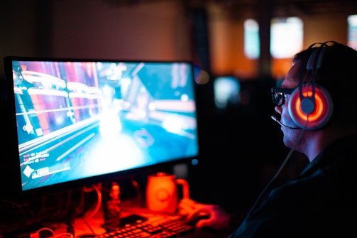 A person wearing headphones is sitting in front of a computer monitor. The screen displays a vibrant, illuminated video game scene, while the person appears focused on gaming. The ambient lighting is dark with red and blue tones, and a backlight from the monitor highlights the headphones.