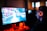 A person wearing headphones is sitting in front of a computer monitor. The screen displays a vibrant, illuminated video game scene, while the person appears focused on gaming. The ambient lighting is dark with red and blue tones, and a backlight from the monitor highlights the headphones.
