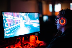 A person wearing headphones is sitting in front of a computer monitor. The screen displays a vibrant, illuminated video game scene, while the person appears focused on gaming. The ambient lighting is dark with red and blue tones, and a backlight from the monitor highlights the headphones.