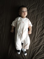 A baby wearing a white onesie is lying on a dark brown blanket. The baby seems calm and is looking slightly upwards. The outfit includes cute animal-themed socks.