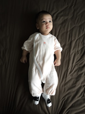 A baby wearing a white onesie is lying on a dark brown blanket. The baby seems calm and is looking slightly upwards. The outfit includes cute animal-themed socks.