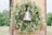 A decorative wreath made of green leaves and small white flowers surrounds a rustic, engraved metal bell, creating a charming and natural look. The wreath is attached to a wooden door panel, suggesting an outdoor or country setting with a blurred green landscape in the background.