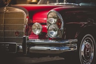 A classic automobile with a deep red paint job and chrome accents. The front grille and rounded headlights are prominent, with the car's emblem visible on the grille. The shiny surface reflects light, giving it a polished, vintage appearance.