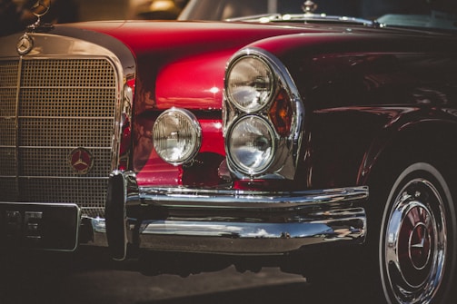 A classic automobile with a deep red paint job and chrome accents. The front grille and rounded headlights are prominent, with the car's emblem visible on the grille. The shiny surface reflects light, giving it a polished, vintage appearance.