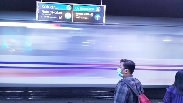 A person wearing a face mask stands on a train platform, with a train blurred in motion passing by. Above, a sign provides directions and information in the Indonesian language, indicating exits and locations such as the airport. The platform is dimly lit, and another person is partially visible on the right side.