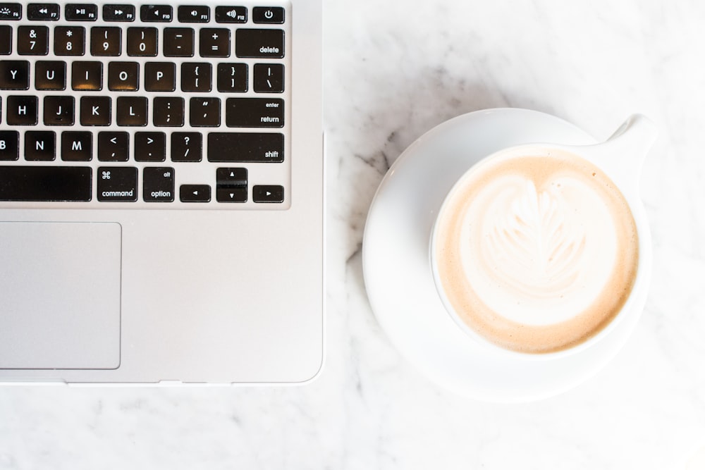 Cappuccino auf Teetasse und Untersetzer neben einem MacBook