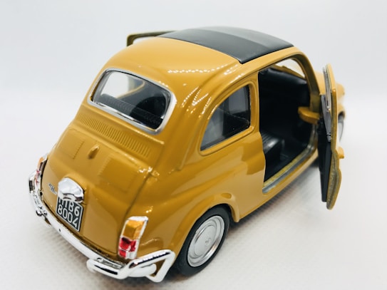 A miniature model of a vintage mustard yellow car with the back door open. The car has a black roof and detailed features including license plate and chrome bumpers. It is positioned on a plain white background.