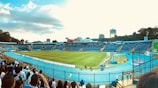 A large stadium with a well-maintained grass football field is surrounded by stands filled with spectators. The seating area is mostly blue, contrasting with yellow accents. In the background, urban buildings and trees are visible under a partly cloudy sky. People can be seen standing near the fence, some wearing sports jerseys.